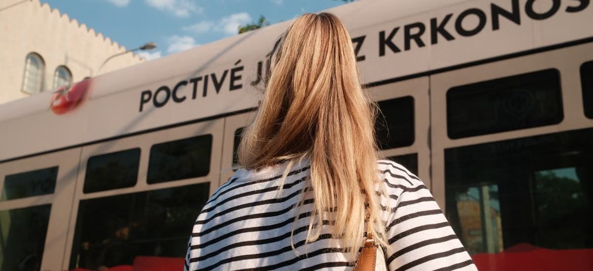 A woman with blonde hair in a striped shirt stands near a city tram on a sunny day.