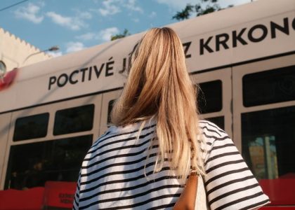 A woman with blonde hair in a striped shirt stands near a city tram on a sunny day.